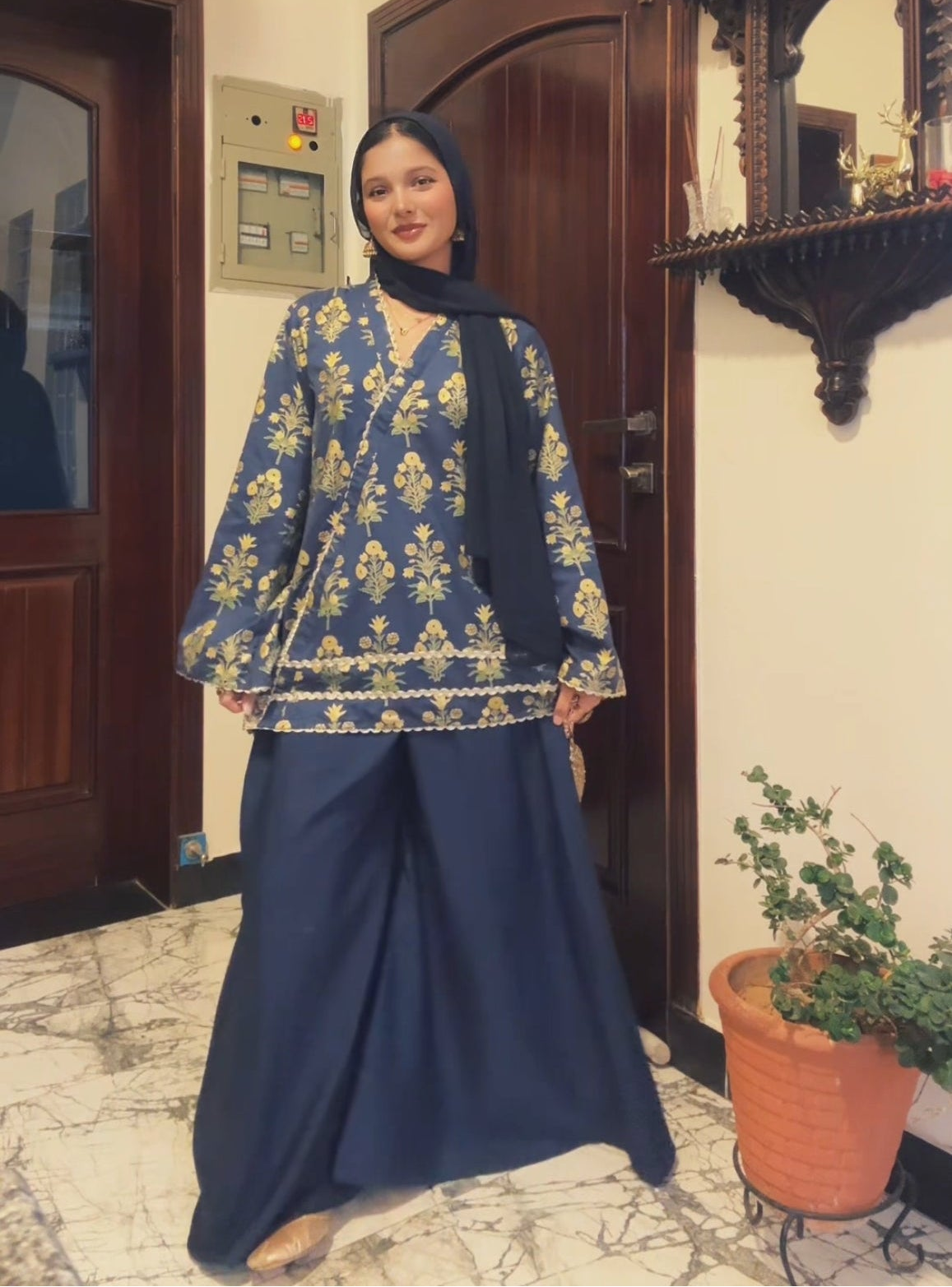 A woman wearing a navy blue traditional outfit with gold floral embroidery and a black hijab, standing in an indoor hallway with wooden doors and a potted plant.